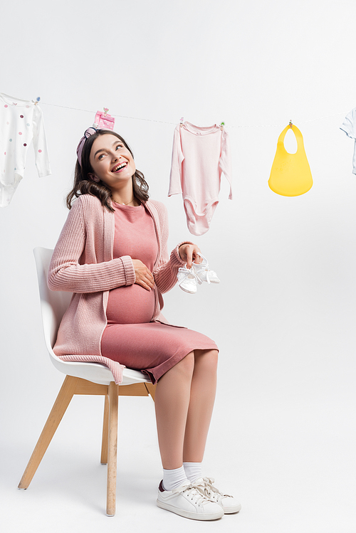 pregnant and pleased woman holding tiny boots and sitting near near baby clothes on clothing line on white