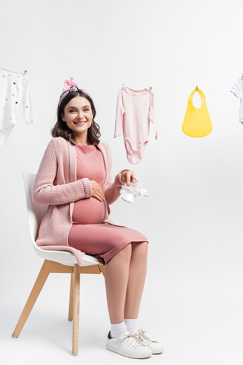 young pregnant woman holding tiny boots and sitting near near baby clothes on clothing line on white