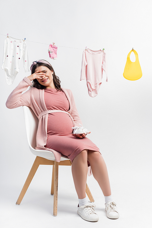 pregnant woman sitting on chair and crying near baby clothes on white