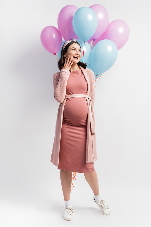 joyful and pregnant woman looking away while standing near balloons on white