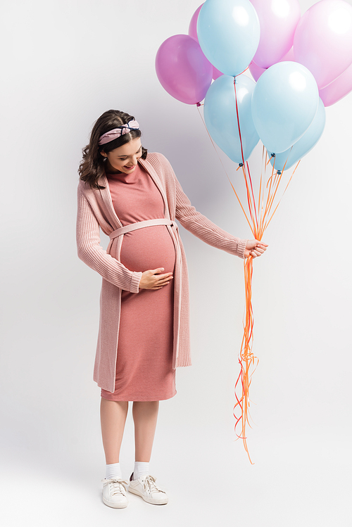 joyful and pregnant woman in dress touching belly and holding balloons on white