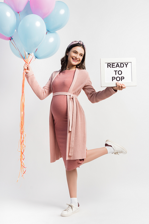 joyful and pregnant woman in dress holding balloons and board with ready to pop lettering on white