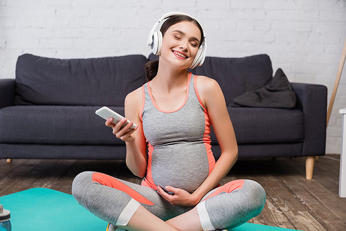 pleased pregnant woman in wireless headphones listening music and holding smartphone