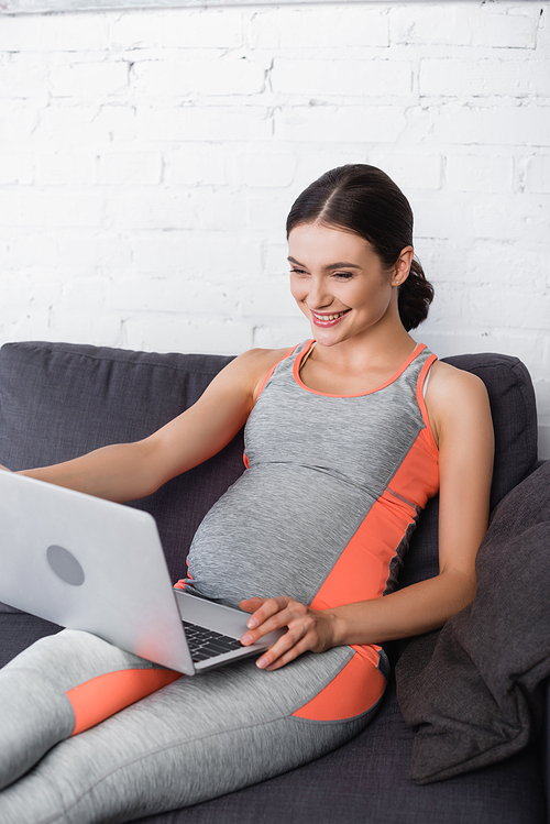 joyful, sportive and pregnant woman looking at laptop in living room