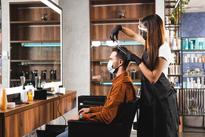 side view of hairstylist in face shield and latex gloves cutting hair of client in medical mask