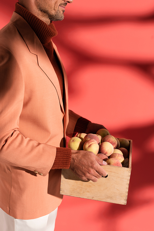 cropped view of happy man in blazer holding box with sweet peaches on red with shadows