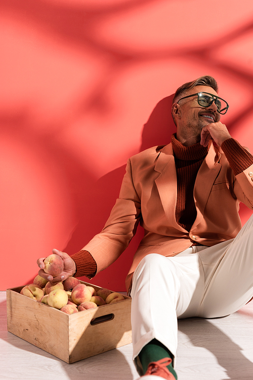 happy man in blazer and sunglasses sitting near box with sweet peaches on red with shadows