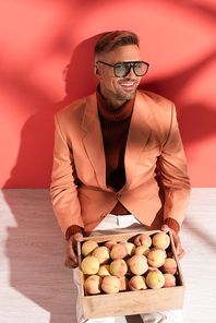 happy man in blazer sitting with sweet peaches in box on red and white with shadows