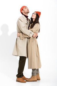 full length of trendy couple in beanie hats and trench coats hugging and looking up on white