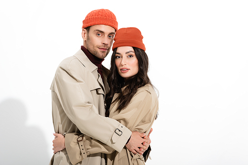 trendy couple in beanie hats and trench coats hugging on white