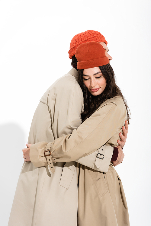 stylish couple in beanie hats and trench coats hugging on white