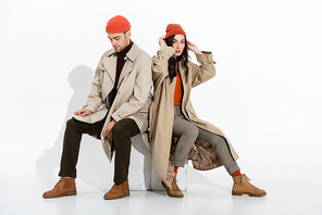 young woman touching beanie hat near trendy man sitting on white