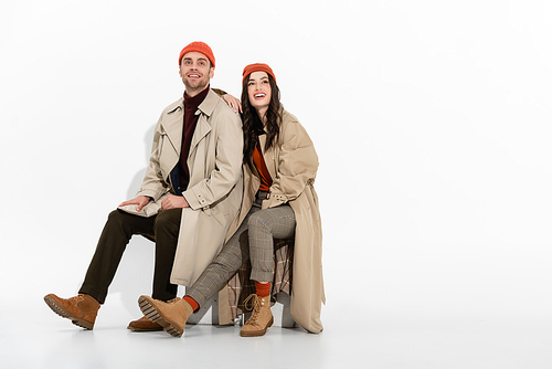 brunette woman in trench coat looking up while sitting near trendy man in beanie hat on white