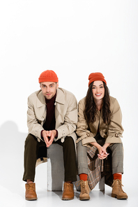 brunette woman in trench coat sitting with clenched hands near trendy man in beanie hat on white