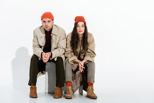 young brunette woman in trench coat sitting with clenched hands near trendy man in beanie hat on white