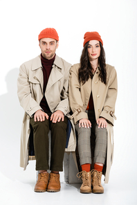 young brunette woman in trench coat sitting near man in beanie hat on white