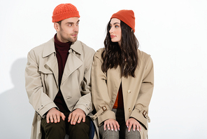trendy couple in beanie hats and trench coats sitting and looking at each other on white