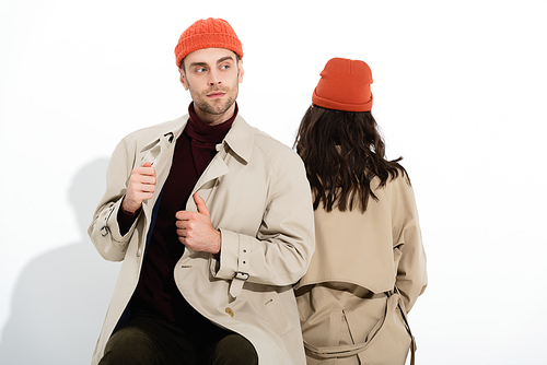 trendy man in beanie hat touching trench coat near woman sitting on white