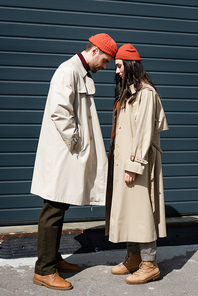 full length of stylish couple in trench coats and hats leaning on each other outside