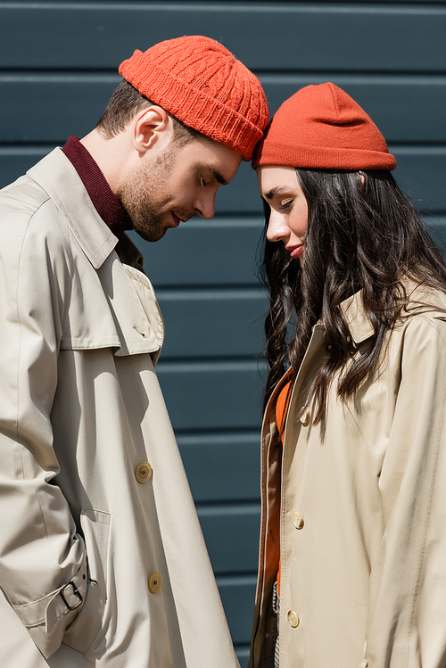 side view of stylish couple in trench coats and hats leaning on each other outside