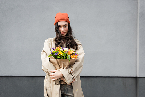 stylish woman in beanie hat and trench coat holding flowers near grey wall