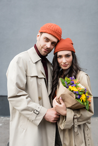 stylish couple in trench coats and hats holding bouquet of flowers