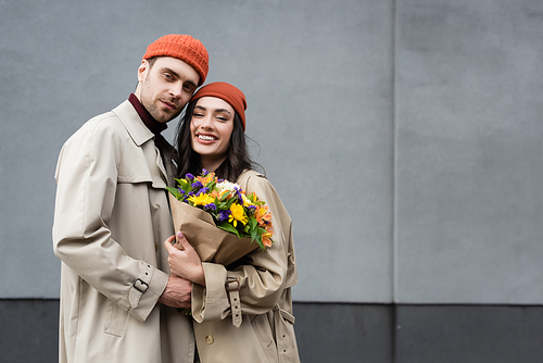trendy couple in trench coats and hats holding bouquet of flowers