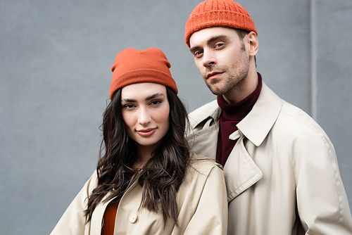 fashionable couple in trench coats and hats standing near grey wall outside
