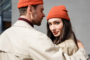stylish man in trench coat hugging woman in beanie hat outside