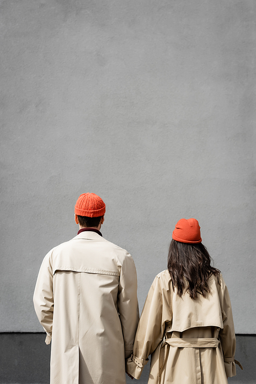back view of couple in trench coats and hats standing near grey wall