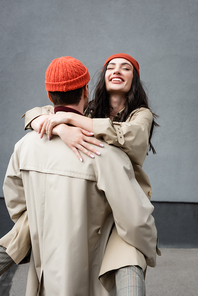 back view of man holding in arms joyful woman in beanie hat and trench coat