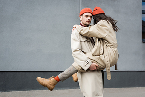 stylish man holding in arms woman in beanie hat and trench coat