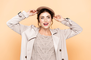 stylish woman in trench coat touching beret while looking away on peach
