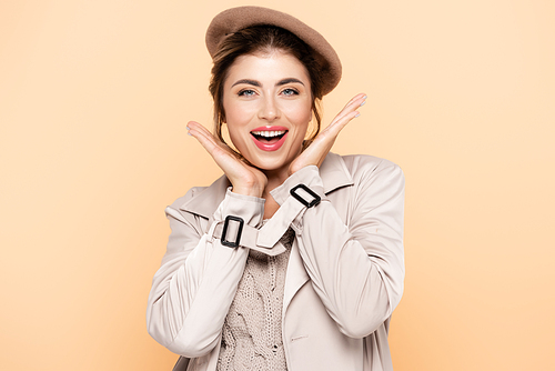 excited woman in beret and trench coat holding hands near face while  isolated on peach