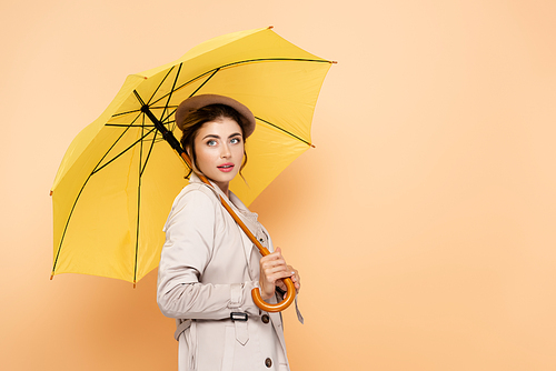 elegant woman in trench coat and beret looking away under yellow umbrella on peach