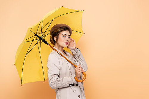 fashionable woman in trench coat and beret touching face under yellow umbrella on peach
