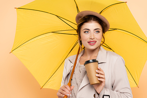 joyful woman in trench coat and beret holding disposable cup under yellow umbrella on peach