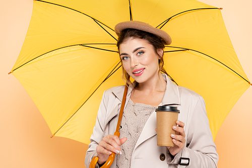 trendy woman in trench coat and beret holding coffee to go under yellow umbrella on peach