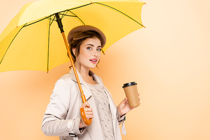 sensual woman in stylish autumn outfit  while holding coffee to go under yellow umbrella on peach