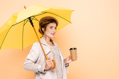 elegant woman in trench coat and beret holding paper cup under yellow umbrella on peach