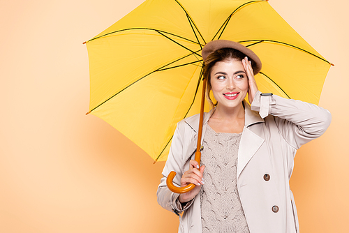 joyful woman in fashionable autumn outfit touching face under yellow umbrella on peach