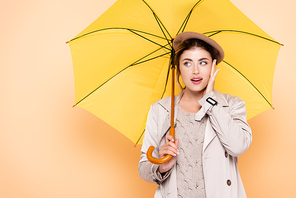 trendy woman in autumn outfit touching face and looking away under yellow umbrella on peach