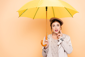 stylish woman in autumn outfit touching face under yellow umbrella on peach
