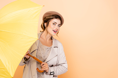 sensual woman in fashionable trench coat and beret  while holding yellow umbrella on peach