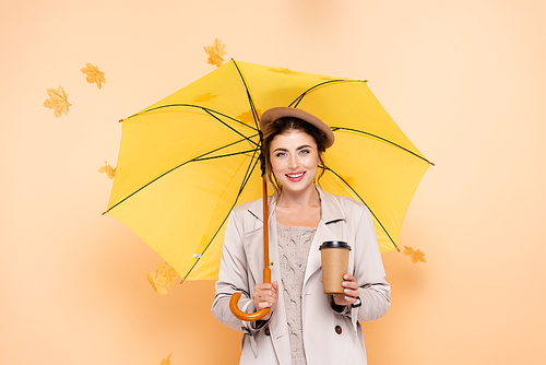 joyful woman in trench coat and beret holding coffee to go under umbrella and falling foliage on peach