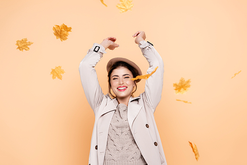 excited woman in trench coat and beret throwing yellow leaves on peach