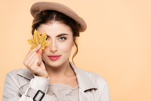 trendy woman in autumn outfit covering eye with yellow leaf while  isolated on peach