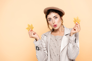 amazed woman in trendy autumn outfit holding yellow leaves isolated on peach