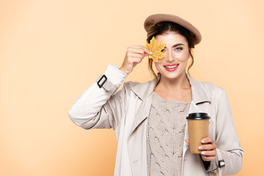 joyful woman in stylish autumn outfit holding coffee to go and covering eye with yellow leaf isolated on peach
