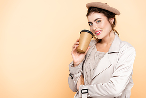 young woman in trench coat and beret holding coffee to go while  isolated on peach